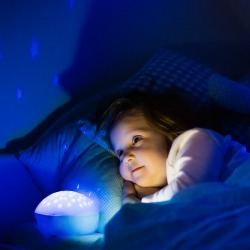 Little girl reading a book in bed. Dark bedroom with night light projecting stars on room ceiling. Kids nursery and bedding. Children read before bedtime. Toddler child playing with lamp and bear toy.